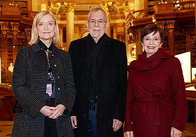 Drei Personen in eleganter Kleidung posieren lächelnd im Marmorsaal. 