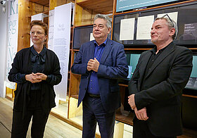 Drei Personen stehen im Literaturmuseum. Hinter ihnen Museumsregale und literarische Texte. 