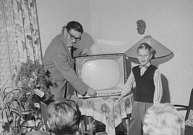 Schwarz-weißes Foto von einer Person im Anzug mit Schnauzer und einem Kind, sie zeigen einen altmodischen Fernseher her, davor sitzen zwei Personen und schauen zu.