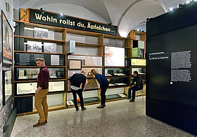 Besucher*innen schauen sich die Ausstellungsregale und Informationstafeln an. 