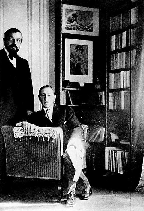 Debussy_Stravinsky_Wohnung.jpg Schwarz-weißes Foto von einem sitzendem und einem stehenden Mann in einer Wohnung mit Bücherregal und ein Kunstdruck von Hokusai an der Wand.