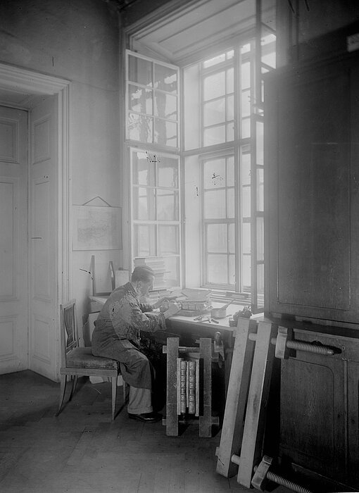 Schwarz-weißes Foto von einem Mann der an einem Schreibtisch vor einem sehr hohen Fenster sitzt