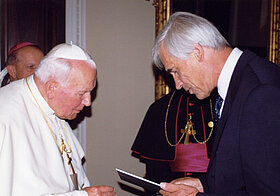 Papst_Johannes_Paul_II.jpg Weißhaarige Person im schwarzen Anzug überreicht Papst Johannes Paul II eine Urkunde.