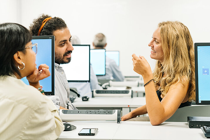 Drei Personen sitzen in einem Computer-Raum und unterhalten sich.