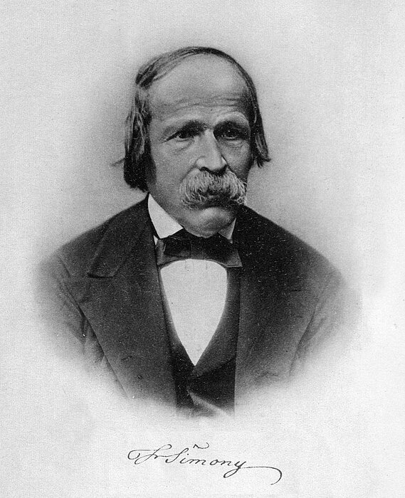 Schwarz-weißes Foto von Person mit Schnauzer und Anzug, darunter steht der Name.