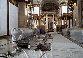 Planen und Gerüste auf dem Boden zu sehen. Rechts hinten stehen 2 Personen.