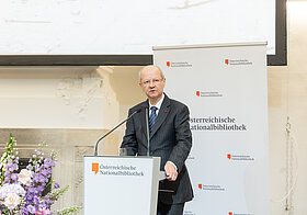 Person in dunklem Anzug hält Rede an Pult, daneben große Blumendekoration.