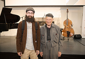 Zwei Personen stehen vor einer Bühne mit Musikinstrumenten und posieren für ein Foto. 