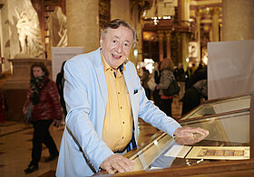 Ältere Person in hellblauem Anzug im Prunksaal.