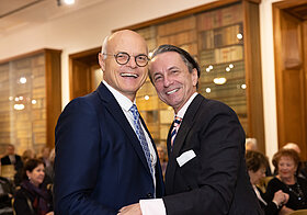Karl_Stoss_Christian_Rainer.jpg Zwei grinsende Personen bei Veranstaltung in Raum mit Bücherregalen.