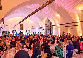 25_galadinner_camineum.jpg Mehrere Personen sitzen in einem großen Saal auf runden Tischen bei einem Gala-Dinner.