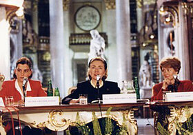 Hillary_Clinton.jpg Drei Personen sitzen an einem Tisch mit Mikrofonen im Prunksaal.