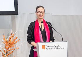 Irmgard_Harrer.jpg Elegant gekleidete Person steht vor einem Rednerpult und spricht.