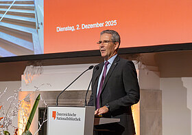 Hartwig_Loeger.jpg Elegant gekleidete Person spricht an einem Rednerpult