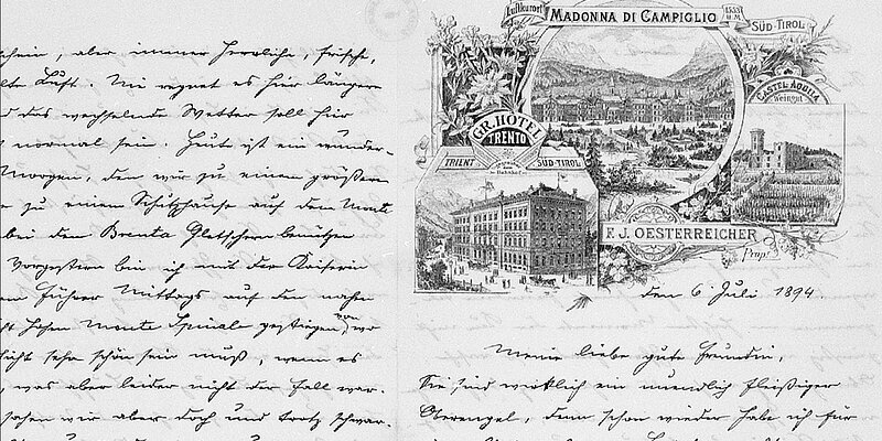 Schwarz-weißes Bild einer handschriftlichen Seite in Kurrentschrift, links dichter Text, rechts eine Illustration mit Landschaftsansicht von Madonna di Campiglio in Südtirol, einem Hotelgebäude und einer Burg, umgeben von dekorativen Pflanzenmotiven. Darunter ein Datum: 6. Juli 1894.