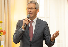 Hartwig_Loeger.jpg Elegant gekleidete Person spricht mit einem Mikrofon.