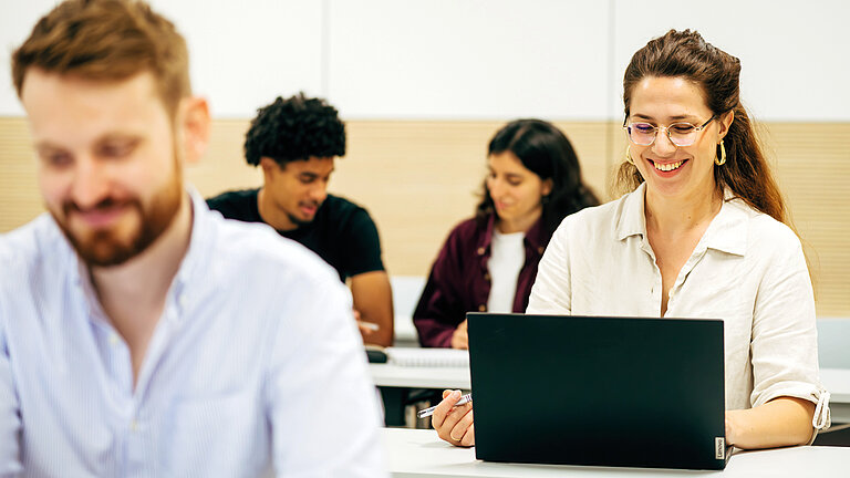 Wissensbisse.jpg Personen sitzen in einem modernen Seminar- oder Klassenraum an Tischen. Im Vordergrund ist eine Person mit einem Laptop zu sehen, im Hintergrund weitere Personen ohne sichtbare Geräte.