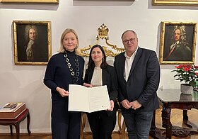 thalhammer_buchpatenschaft.jpg Drei Personen halten Urkunde, im Hintergrund hängen alte Gemälde in Goldrahmen.