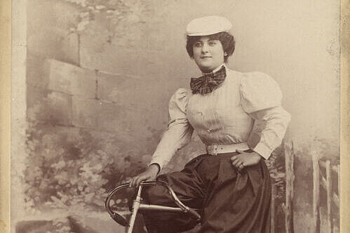 Abb.00Teaserbild.jpg Sepia-Foto von einer Person mit Rock, Bluse und Kappe auf einem Fahrrad