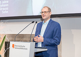 Marko_Deisinger.jpg Elegant gekleidete Person spricht an einem Rednerpult.
