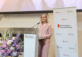 Johanna_Rachinger.jpg Person in rosa Anzug hält eine Rede an einem Pult, daneben große Blumendekoration.