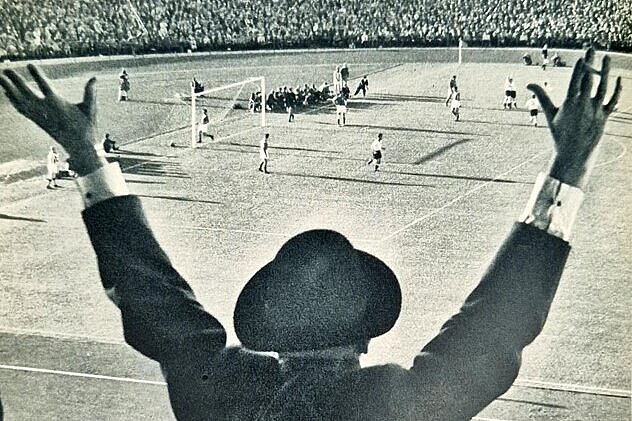 Schwarz-weißes Foto von einer Person von hinten, die die Arme in die Lüfte wirft, vor der Person ein Fußballspiel.